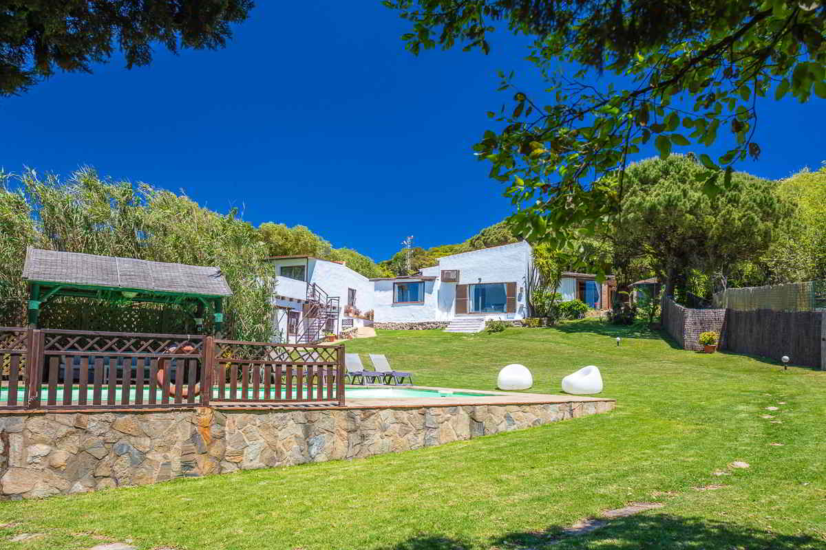 Vista del jardín del Chalet La Ahumada con piscina vallada, pérgola de sombra, tumbonas y fachada blanca del edificio principal al fondo.