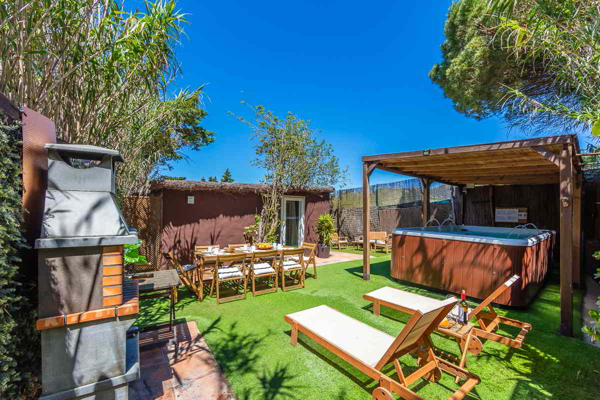 Vista del jardín privado de la Villa Verde en Tarifa, con césped artificial, tumbonas de madera, una mesa grande de comedor al aire libre, barbacoa de obra y un jacuzzi bajo pérgola, todo rodeado de vegetación natural.